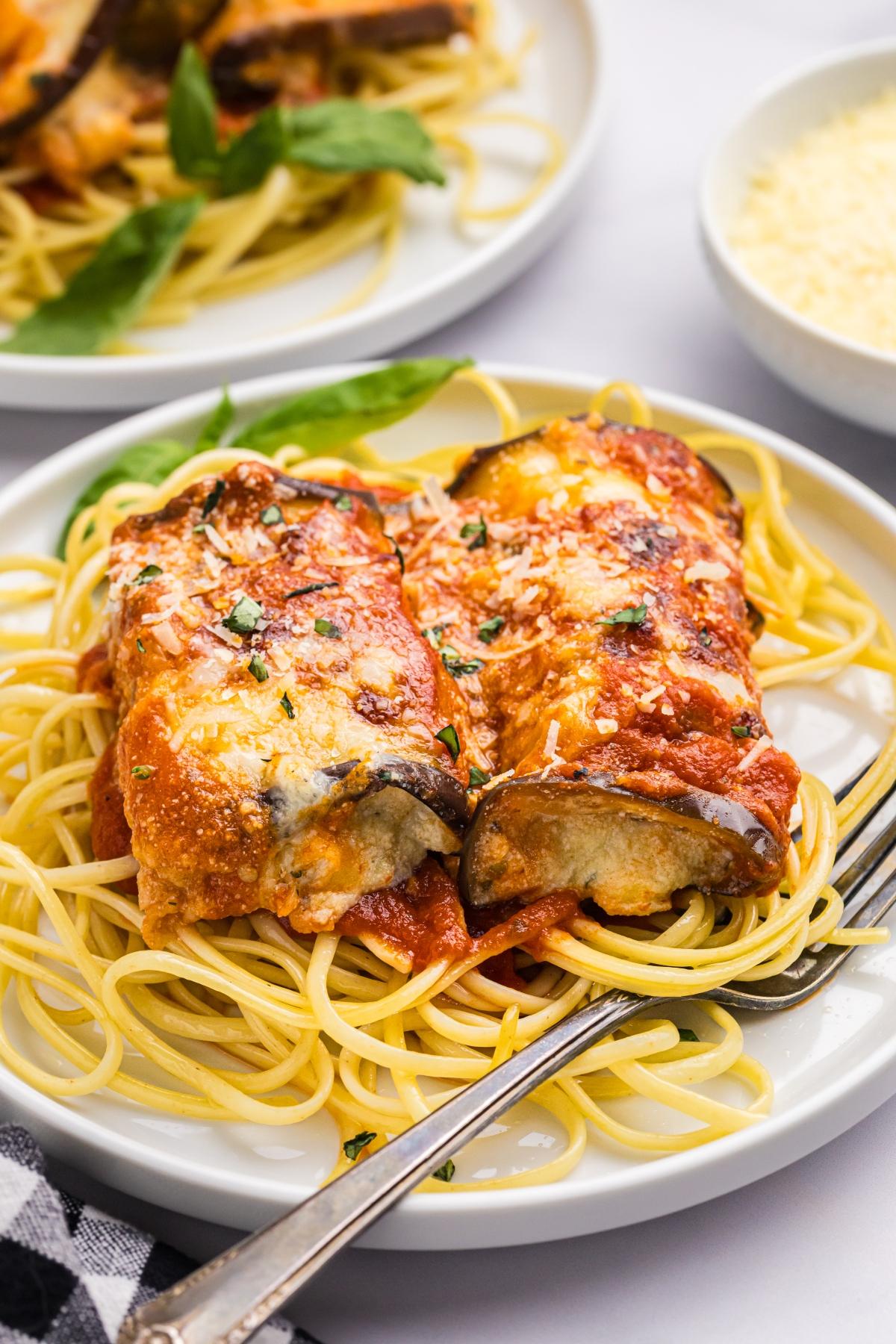 Two pieces of eggplant rollatini on spaghetti, topped with tomato sauce and cheese, served on a white plate.