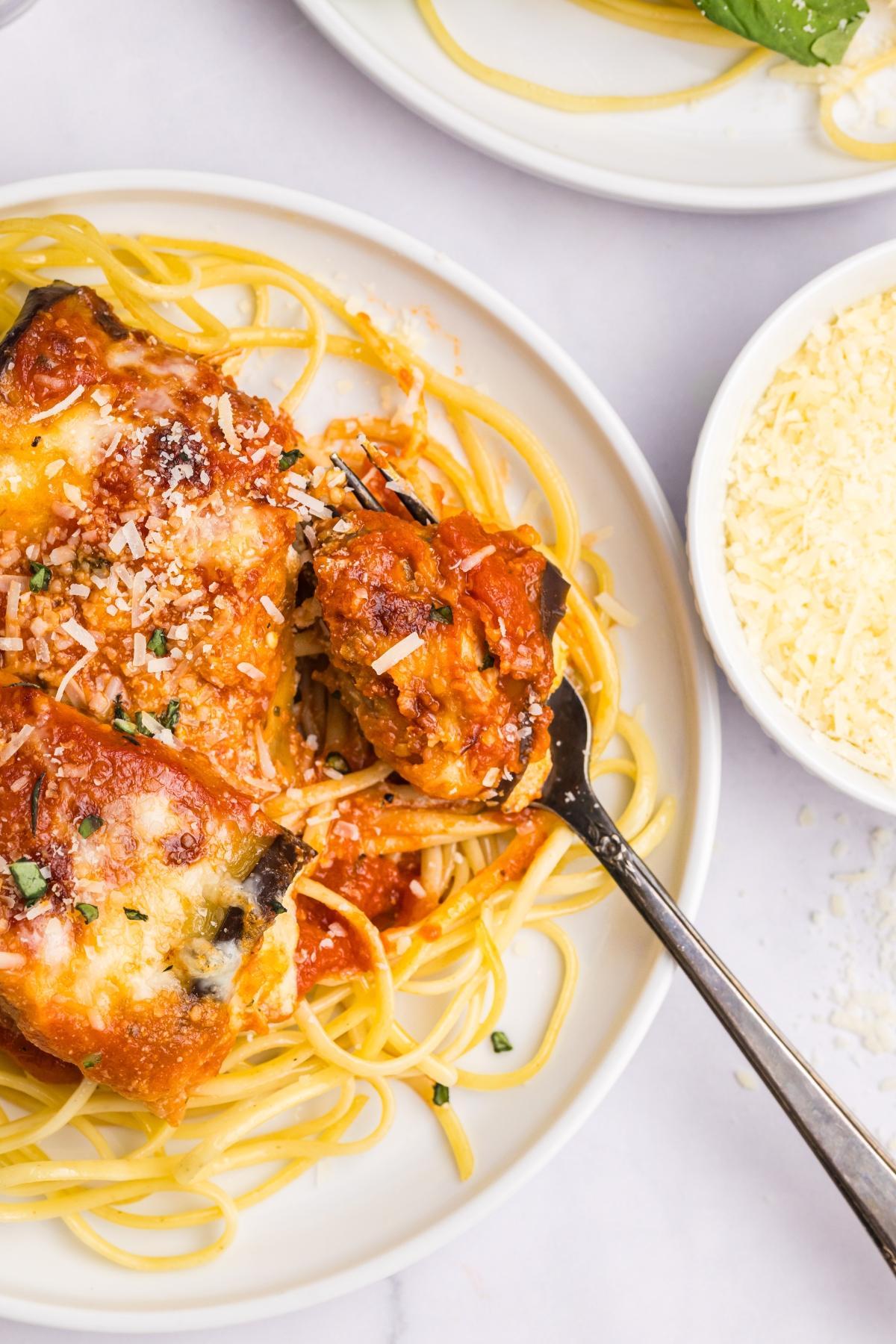 Plate of spaghetti topped with cheesy baked eggplant rollatini, with a fork and a bowl of grated cheese nearby.