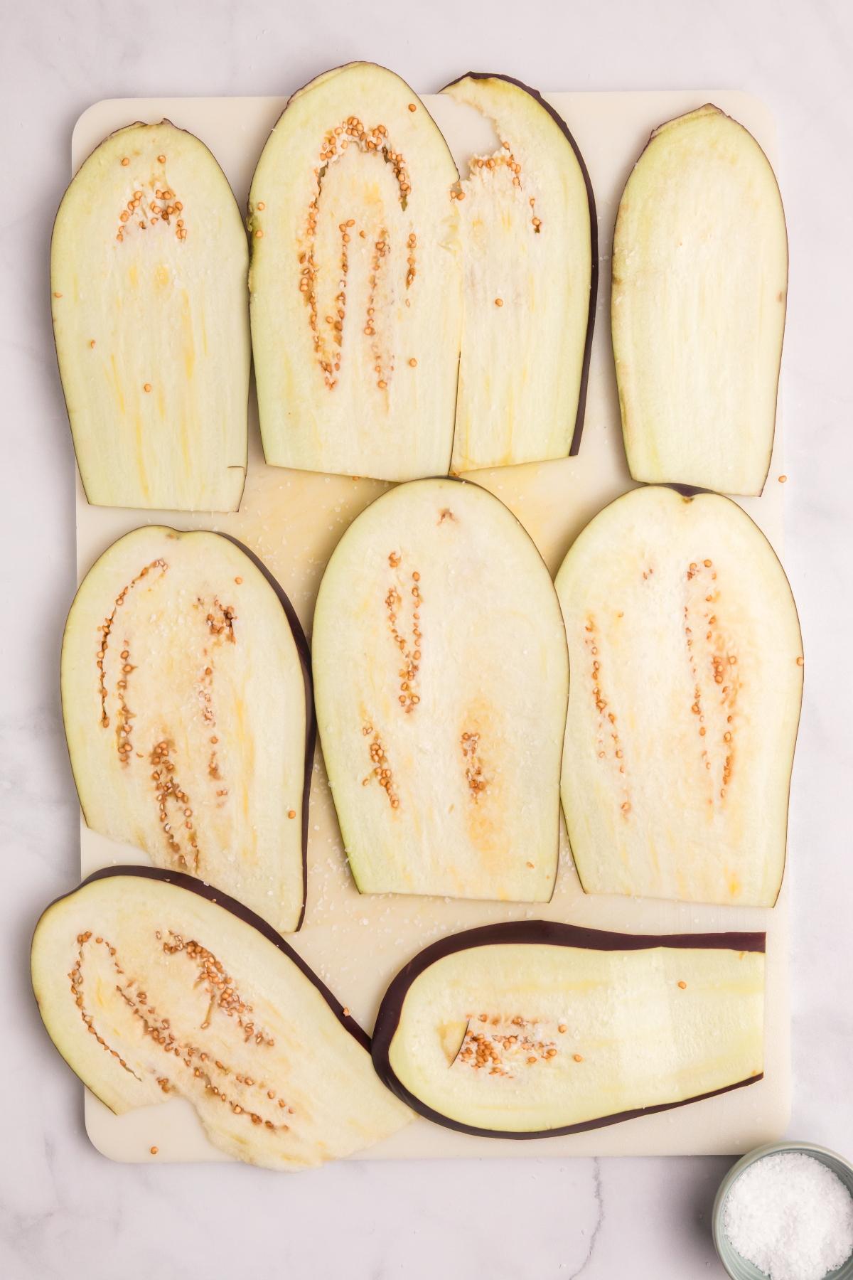 Nine raw eggplant slices are arranged on a white cutting board, with a small bowl of salt nearby.