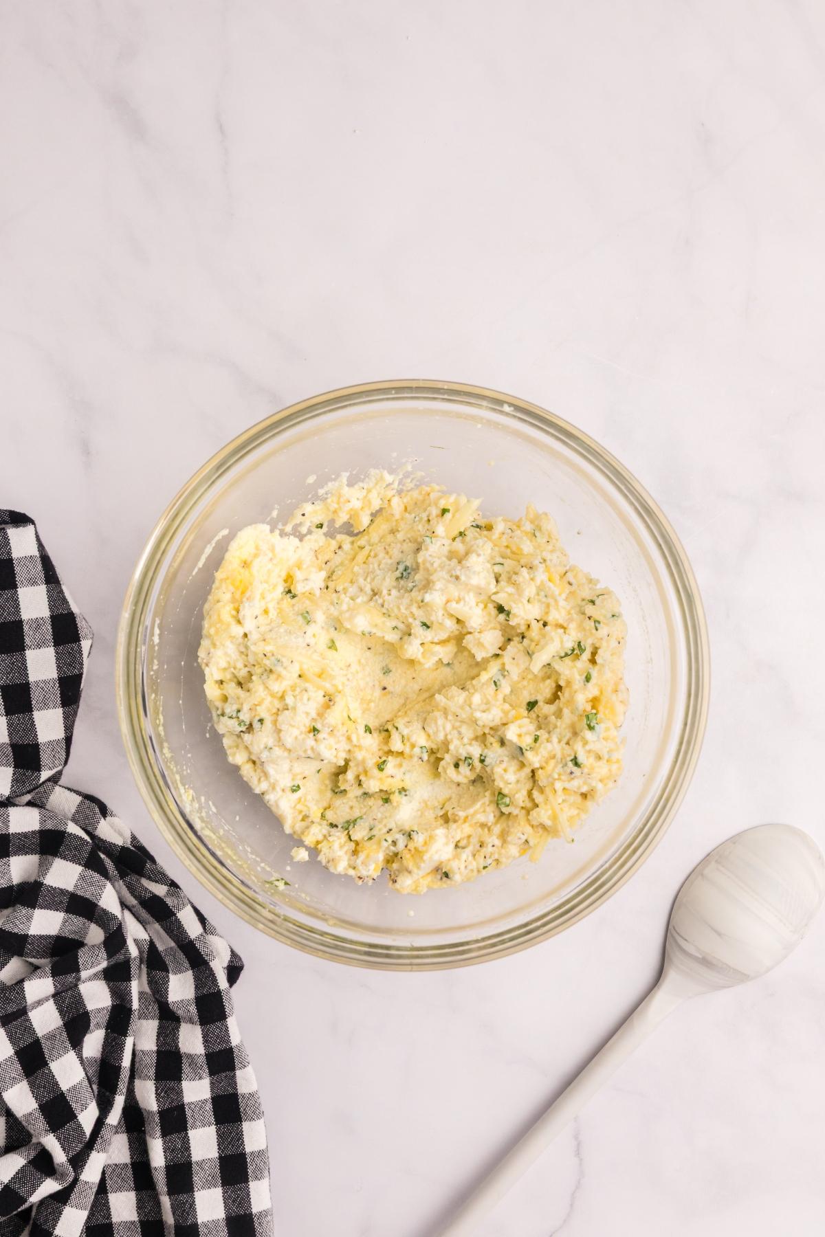 A glass bowl of ricotta cheese mixture beside a black and white checkered cloth and a white spoon.