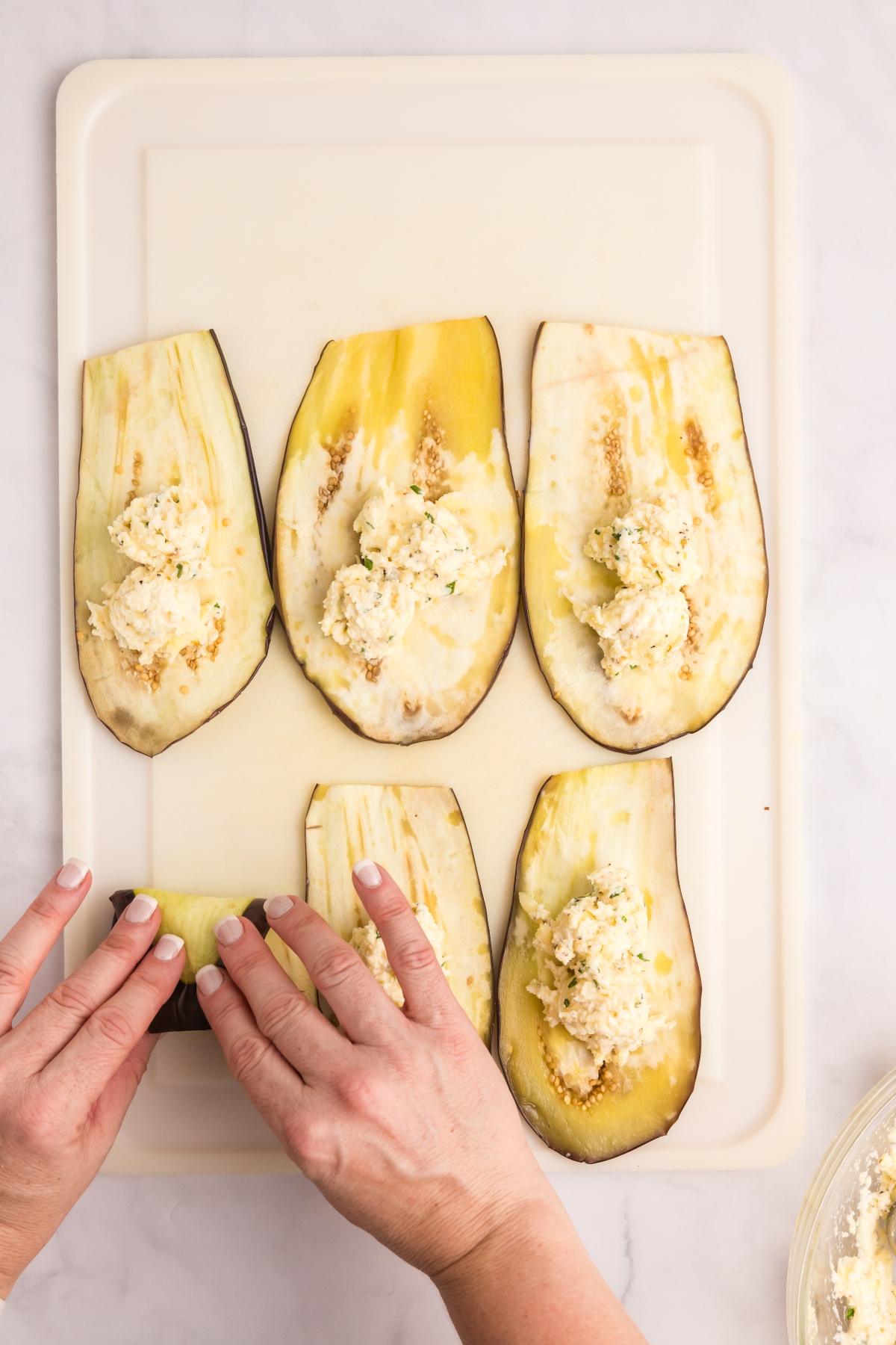 Hands rolling ricotta-filled eggplant slices on a white cutting board, preparing a stuffed dish.