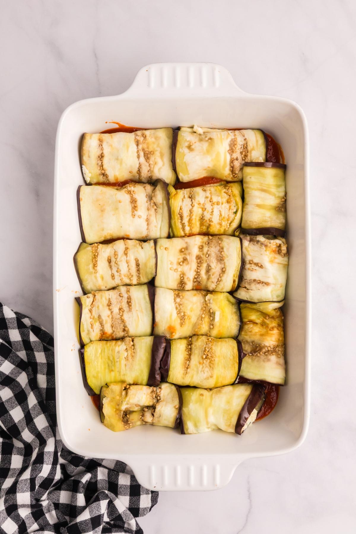 Eggplant rollatini neatly arranged in a white baking dish, next to a black and white checkered towel.