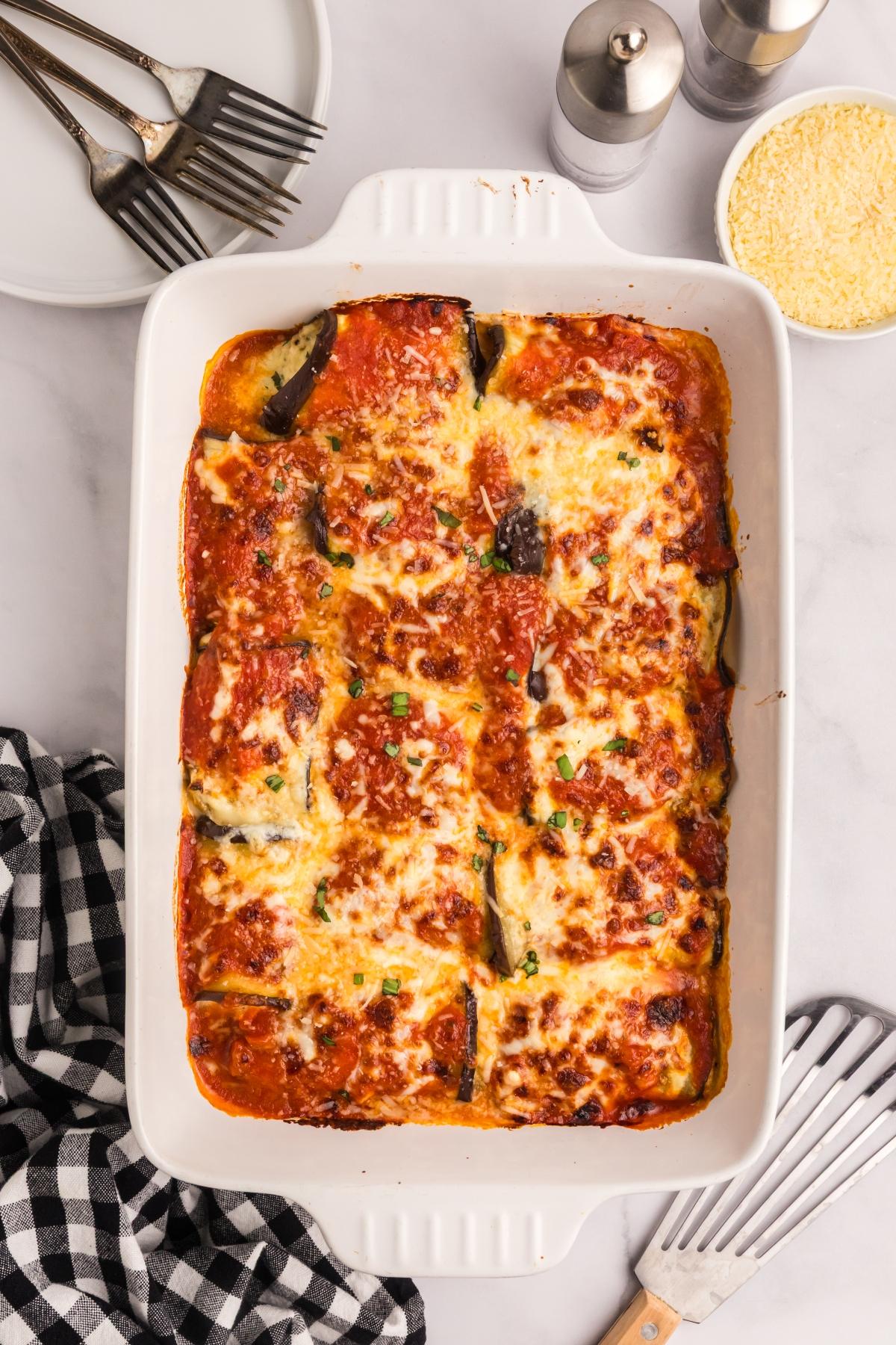 Baked eggplant rollatini in a white dish, with plates, forks, cheese, and seasonings on the side.