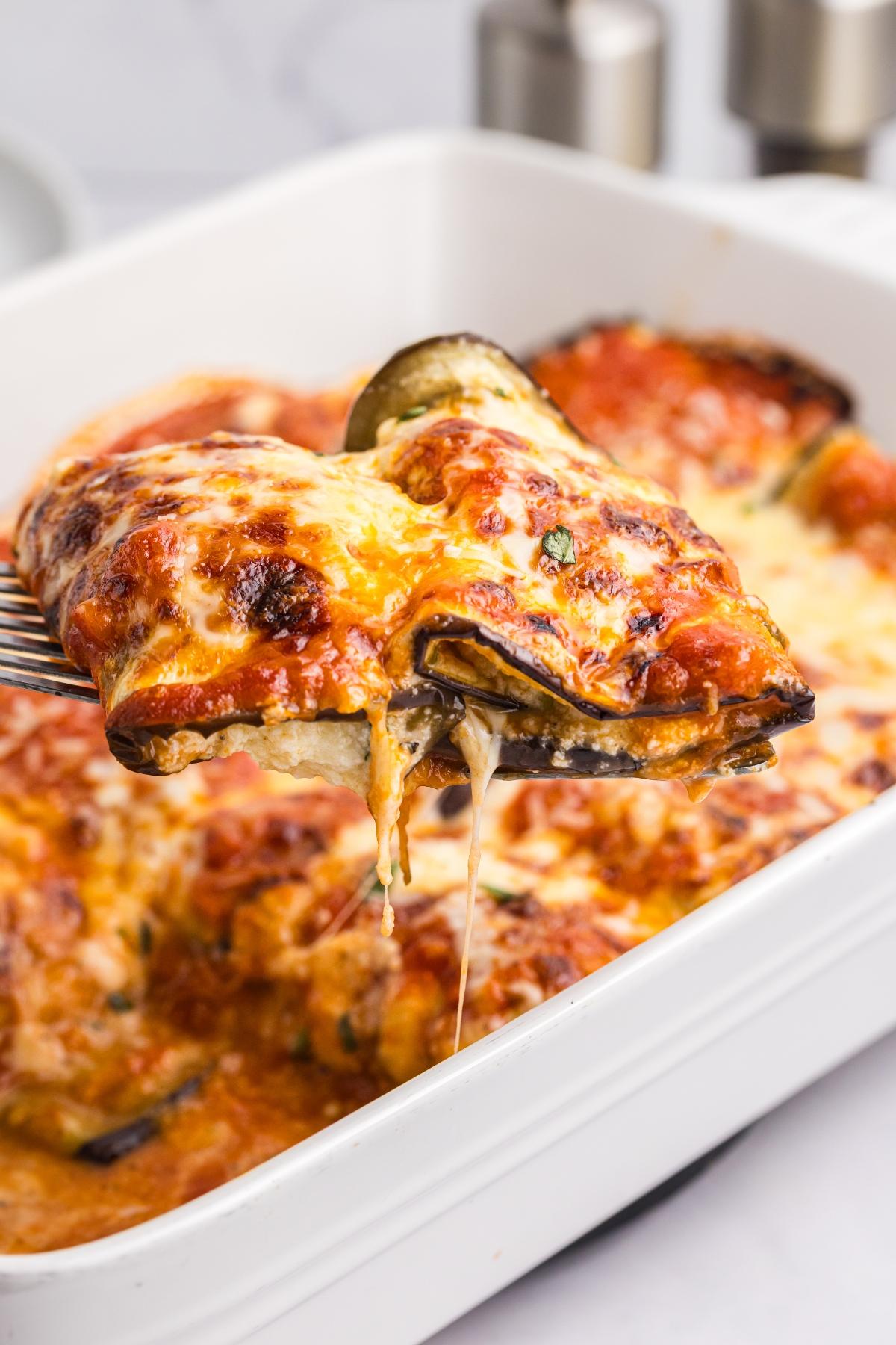 A cheesy baked eggplant rollatini slice being lifted from a white casserole dish.