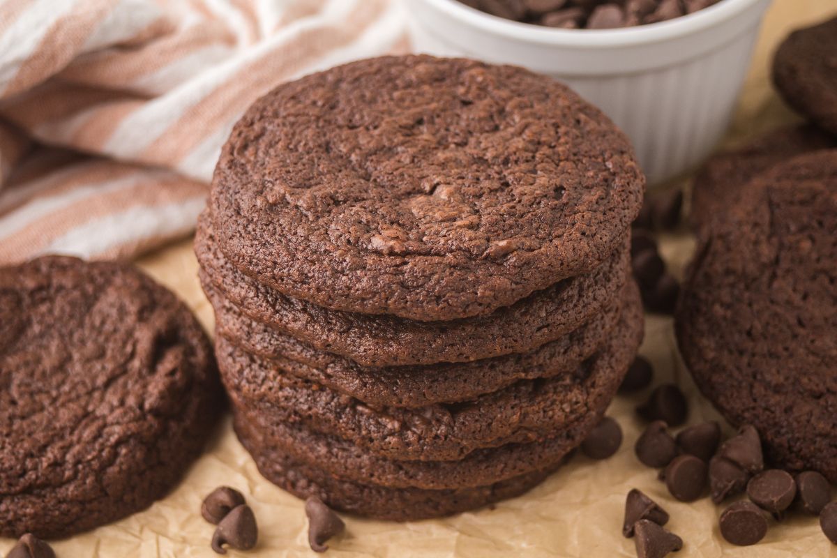A stack of chocolate cookies, reminiscent of espresso brownie cookies, is surrounded by chocolate chips on parchment paper.