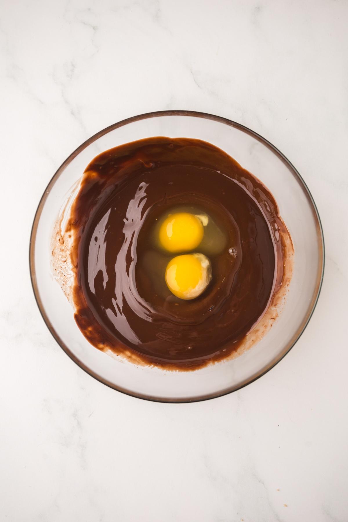 A glass bowl with melted chocolate and two raw eggs on a white marble surface.