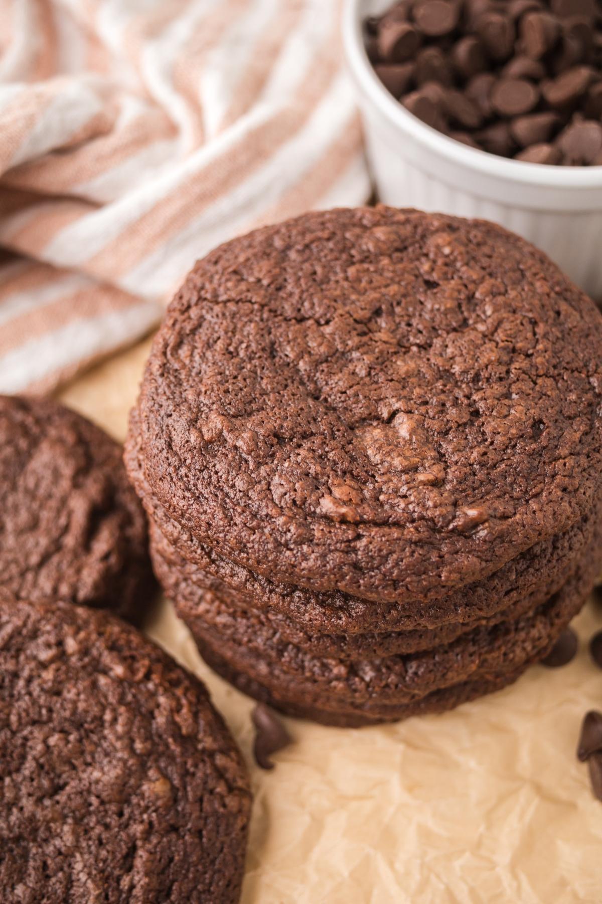 A stack of chocolate cookies next to a bowl of chocolate chips on parchment paper.