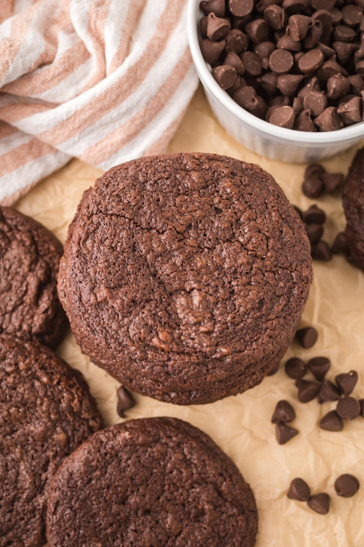 A stack of chocolate cookies next to chocolate chips and a striped towel on parchment paper.