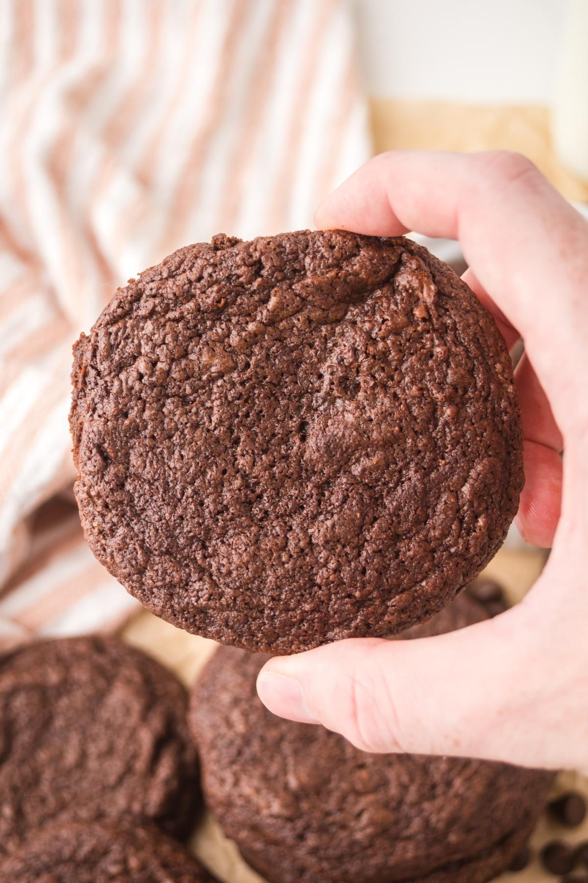 A hand holding a large chocolate cookie above a stack of cookies on a striped cloth background.