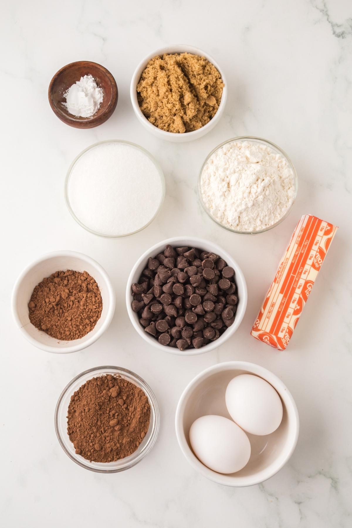 Baking ingredients on a white counter: eggs, butter, chocolate chips, flour, sugars, cocoa, and baking powder.