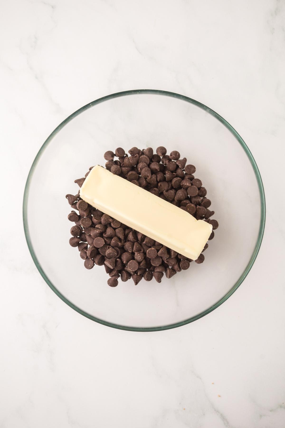A glass bowl with chocolate chips and a stick of butter on a white marble surface.