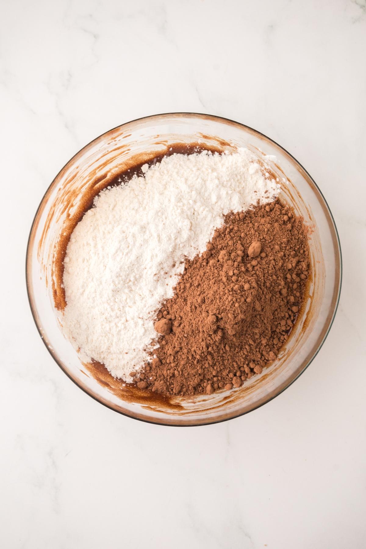 A glass bowl with flour and cocoa powder on a white marble surface, ready to be mixed.