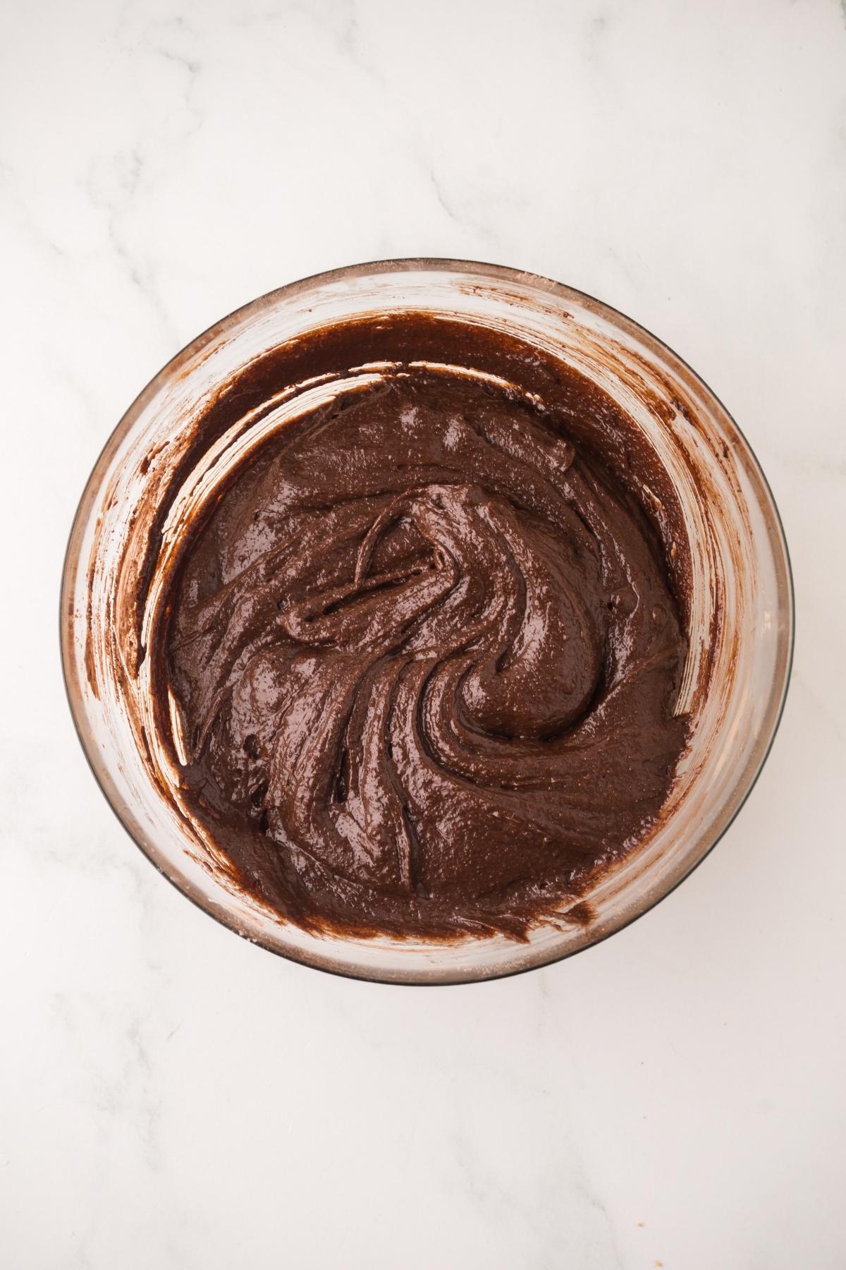 A glass bowl filled with chocolate brownie batter on a white marble surface.