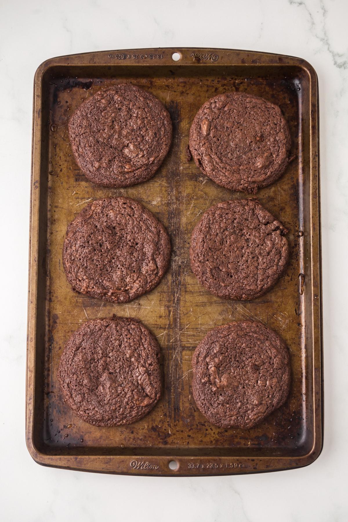 Six large chocolate cookies on a well-used baking sheet, viewed from above on a white surface.