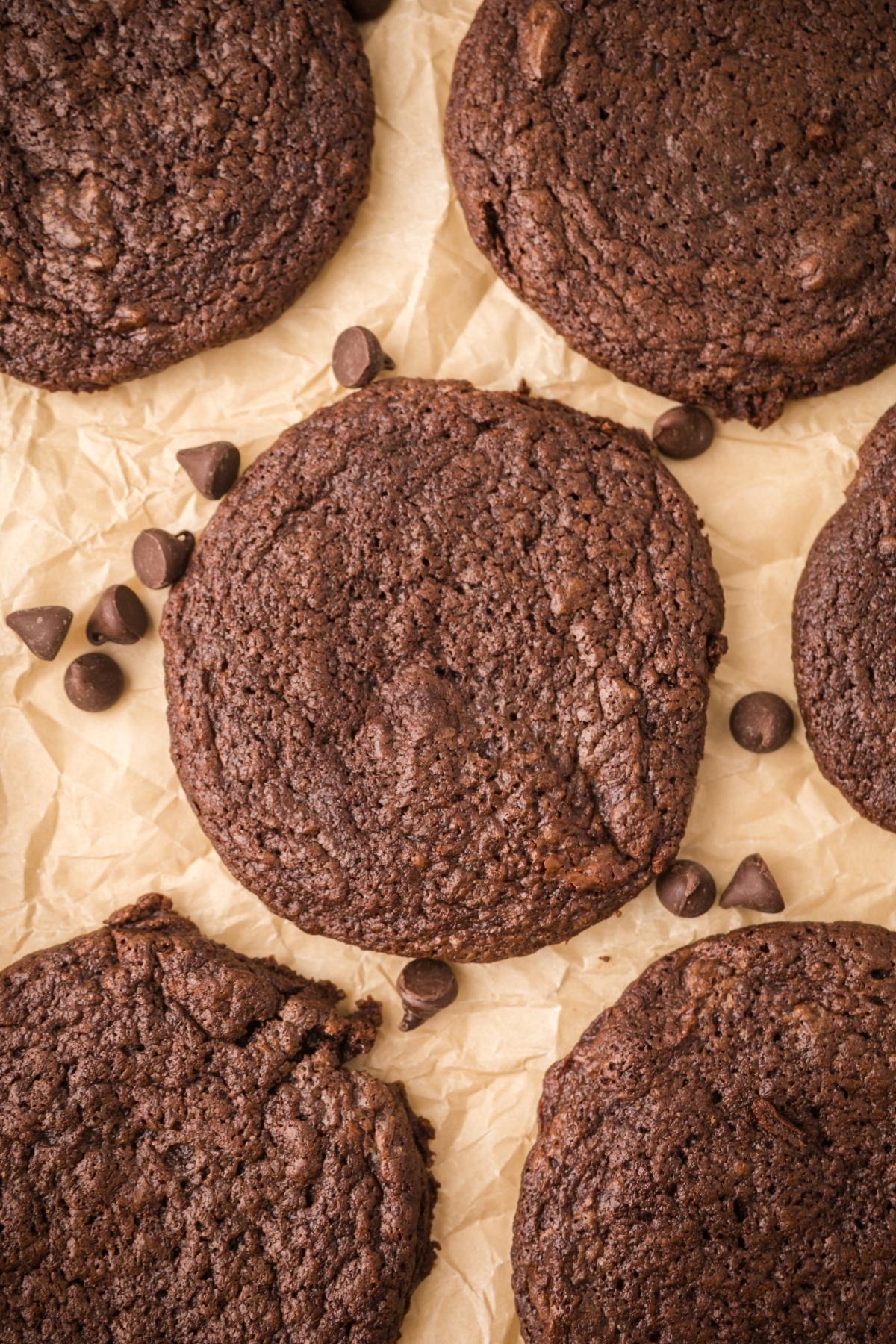 Six double chocolate cookies on parchment paper with a few chocolate chips scattered around.