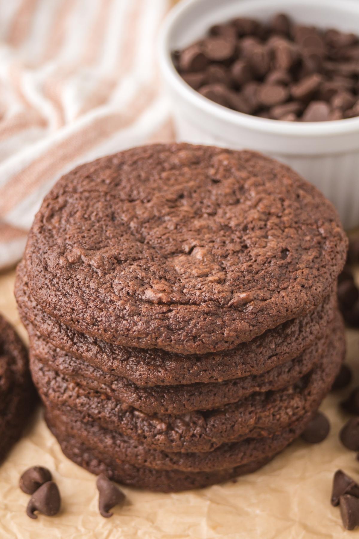 A stack of chocolate cookies sits on parchment paper, with chocolate chips and a bowl of chips nearby.