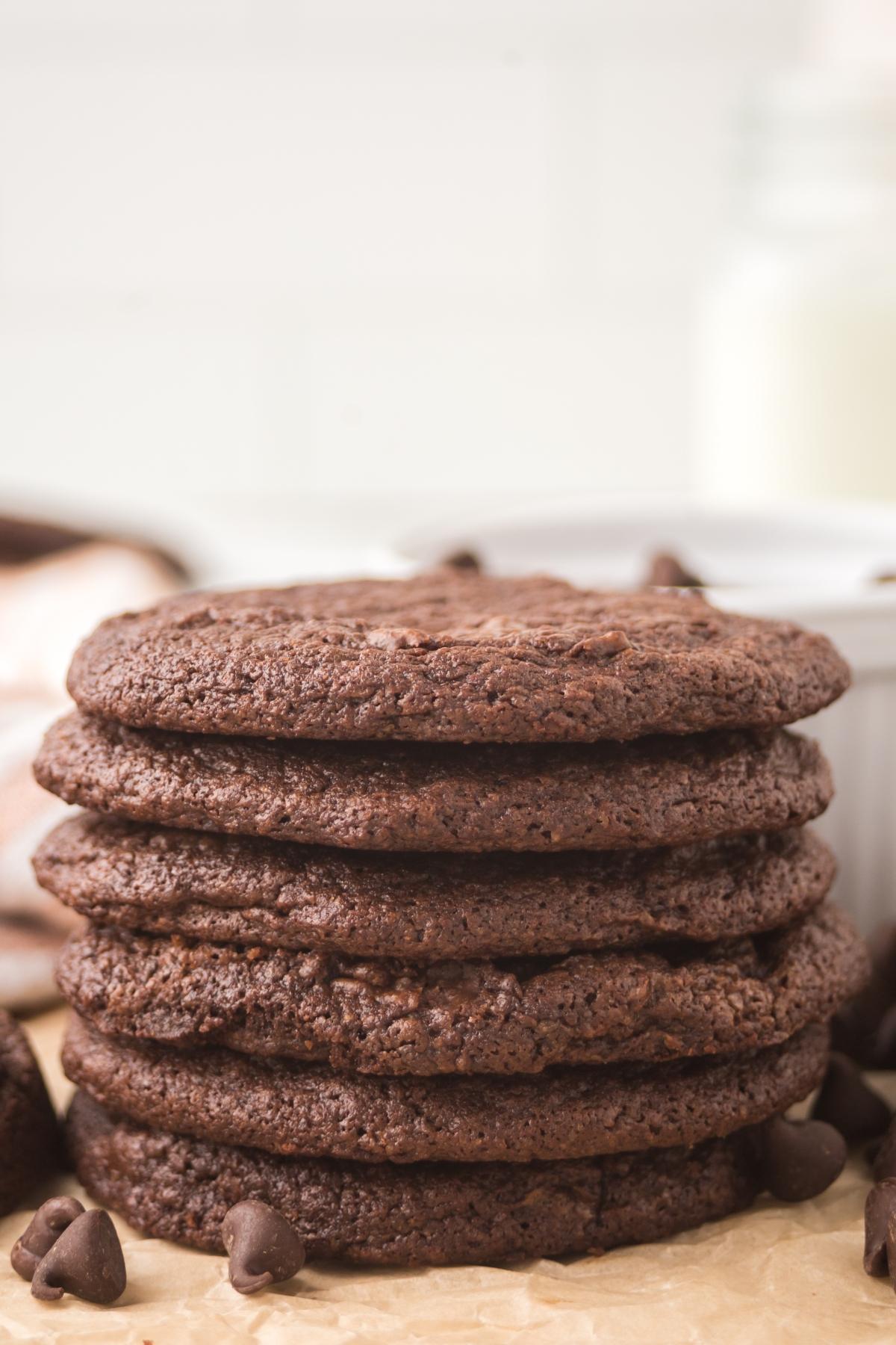 Stack of six chocolate cookies with chocolate chips, on parchment paper, with milk in the background.