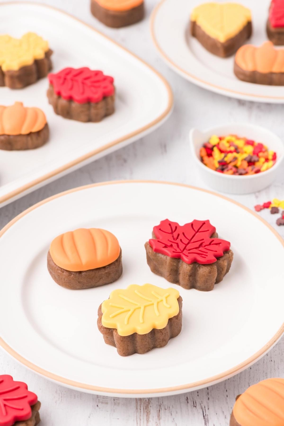 Three fall-themed cake bites, shaped like a pumpkin and leaves, on a white plate with more cookies in the background.