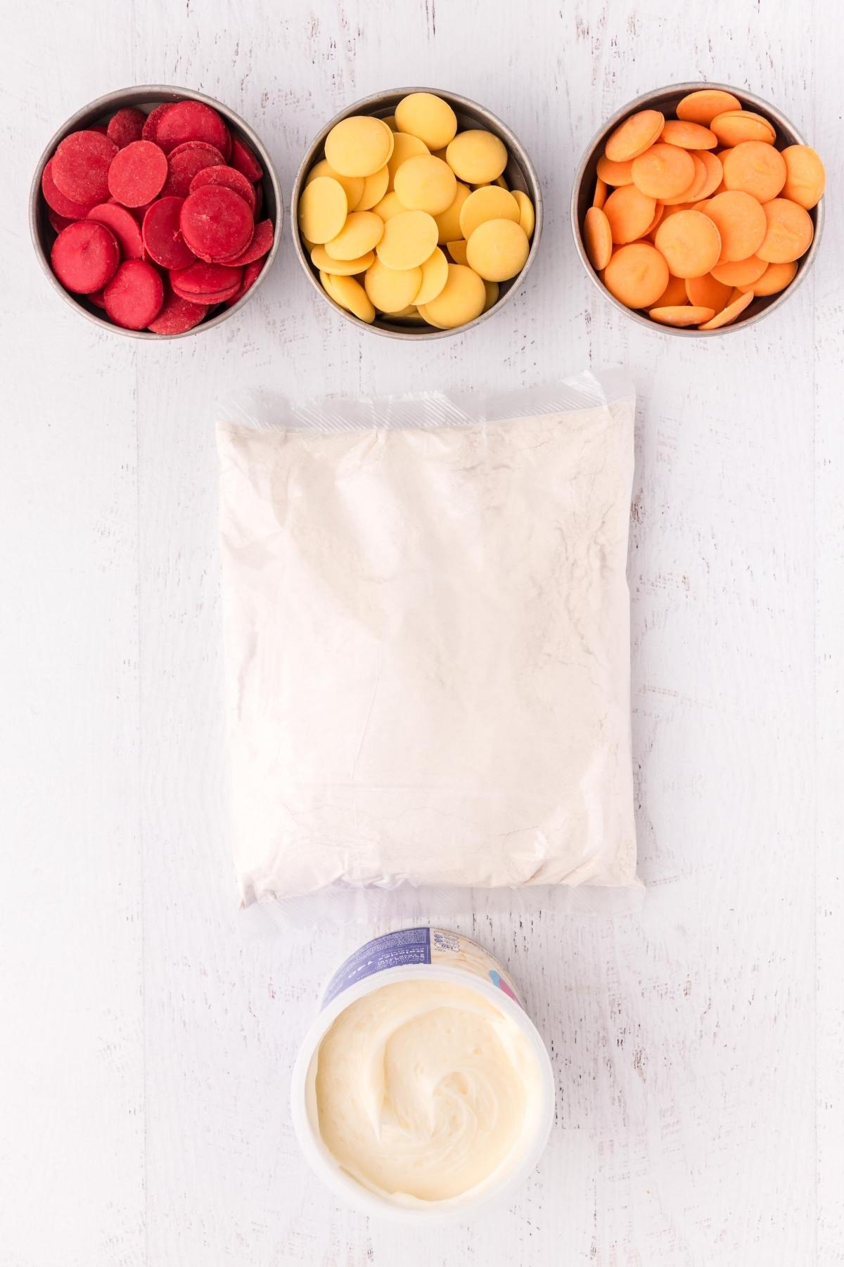 Three bowls of colored candy melts, a bag of powdered mix, and a tub of white frosting on a white surface.