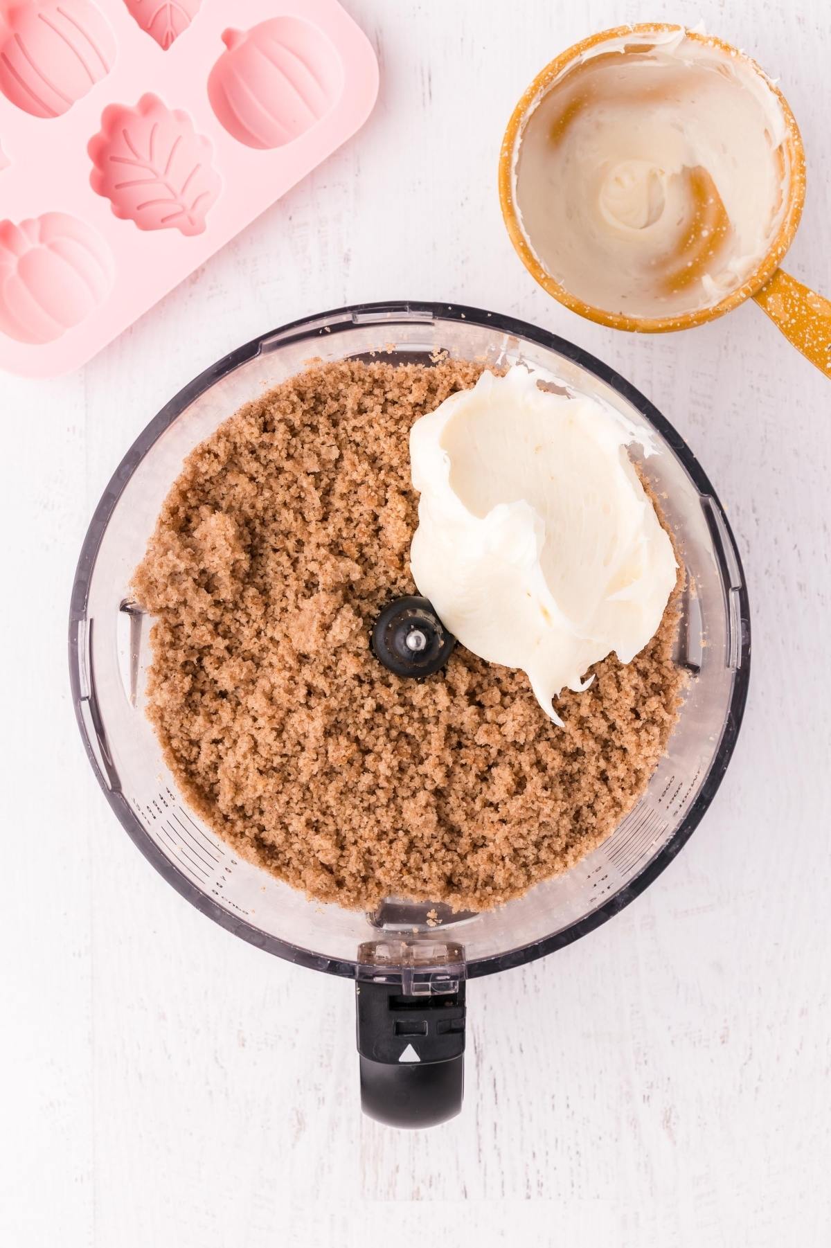 Food processor with brown sugar and cream cheese, and a spatula and pink silicone mold nearby on a white surface.