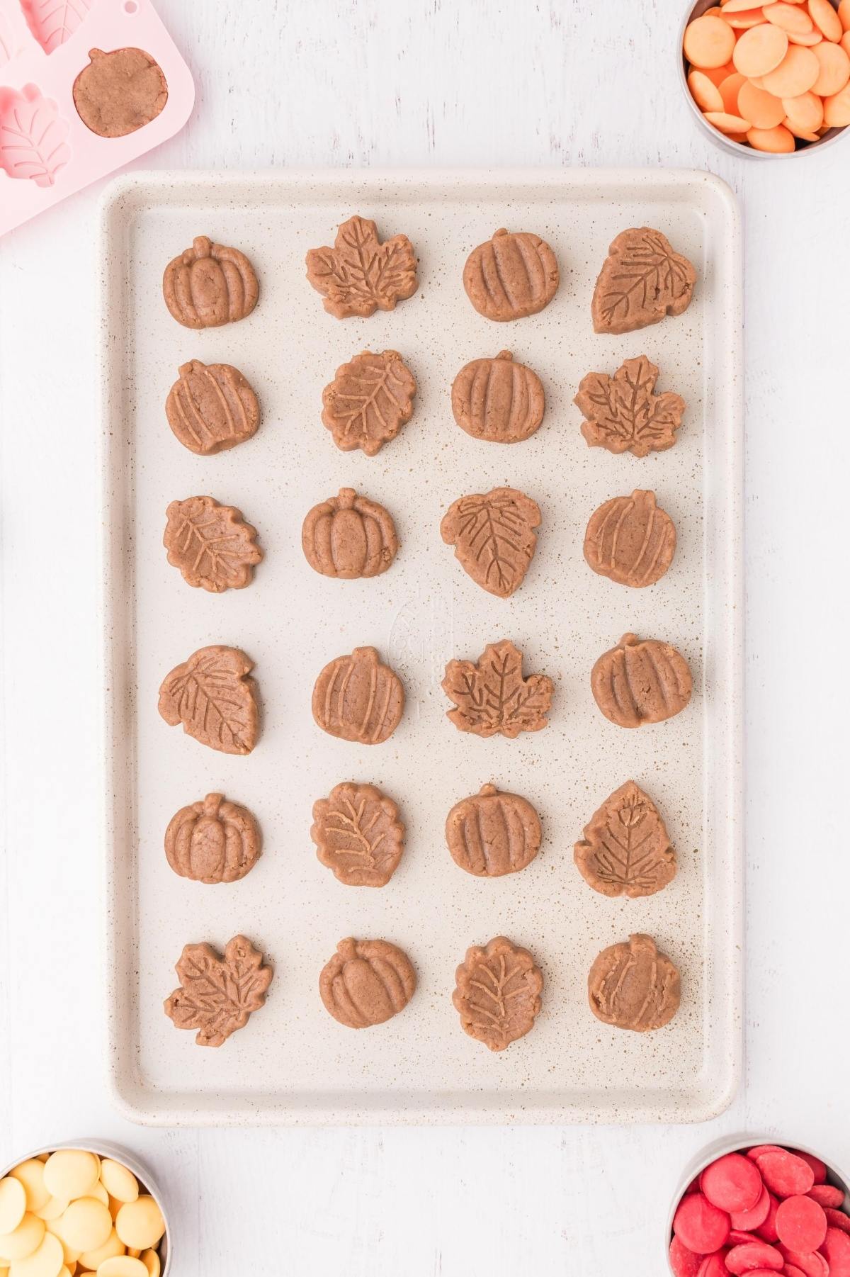 A baking tray with pumpkin and leaf-shaped cookies, surrounded by bowls of colorful candy melts.