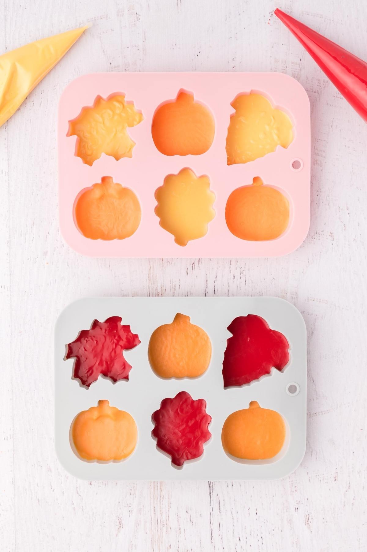 Two silicone molds with fall shapes filled with red and orange batter on a white surface.