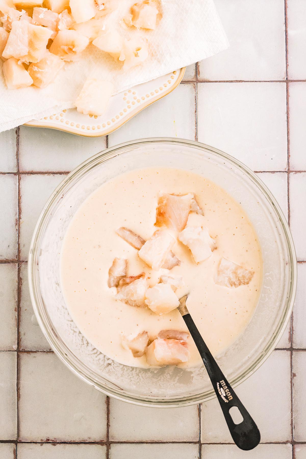 A bowl of batter with fish pieces for fritto misto and a spoon sits next to a plate of more fish on a tiled surface.