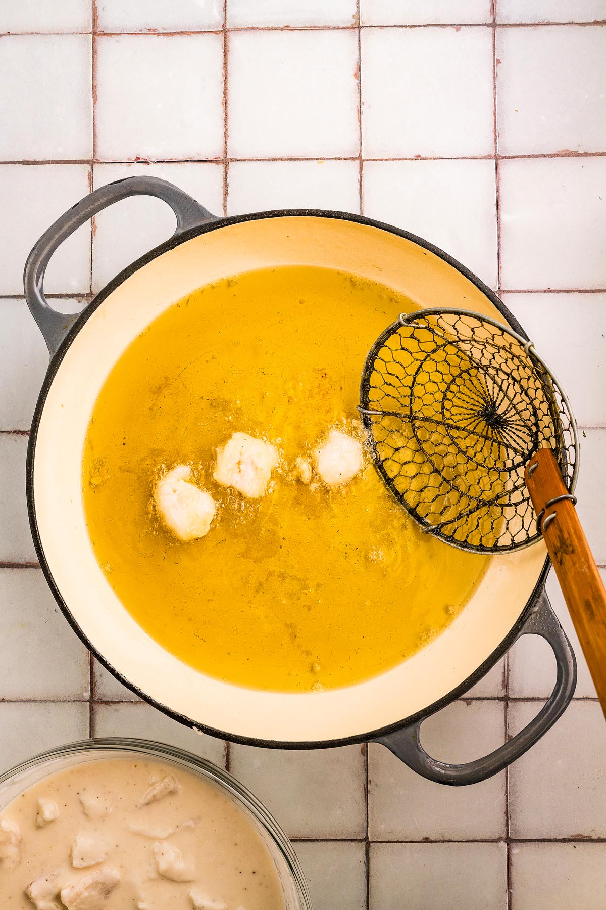 Round pieces of battered food fry in hot oil in a pot; a skimmer rests on the pot&rsquo;s side, ready to lift the golden fritto misto.