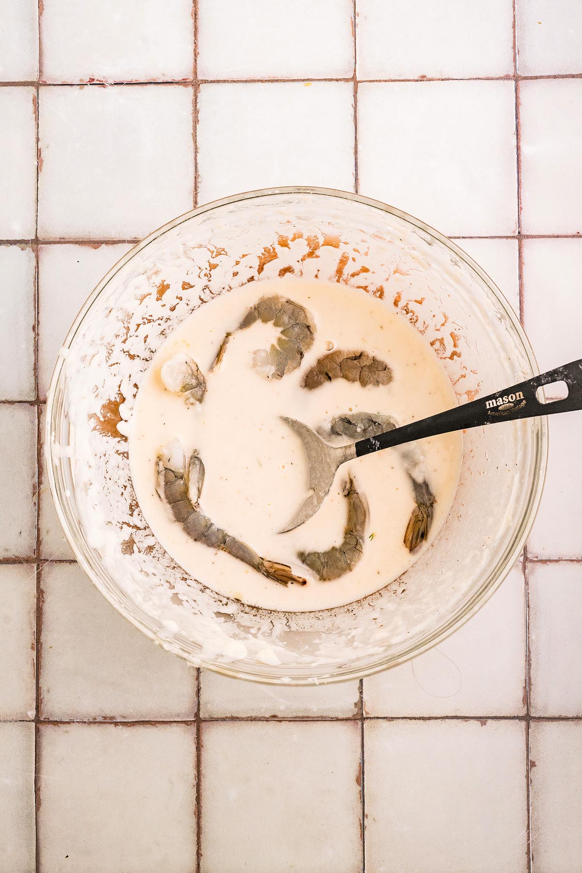 Raw shrimp in a creamy marinade sit in a glass bowl with a black spoon, ready to be the star of your next fritto misto on a white tiled surface.