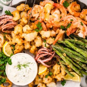 A platter of fritto misto featuring fried seafood, shrimp, green beans, lemon wedges, and dipping sauce, garnished with parsley.