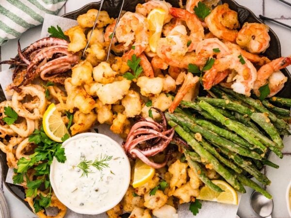 A platter of fritto misto featuring fried seafood, shrimp, green beans, lemon wedges, and dipping sauce, garnished with parsley.