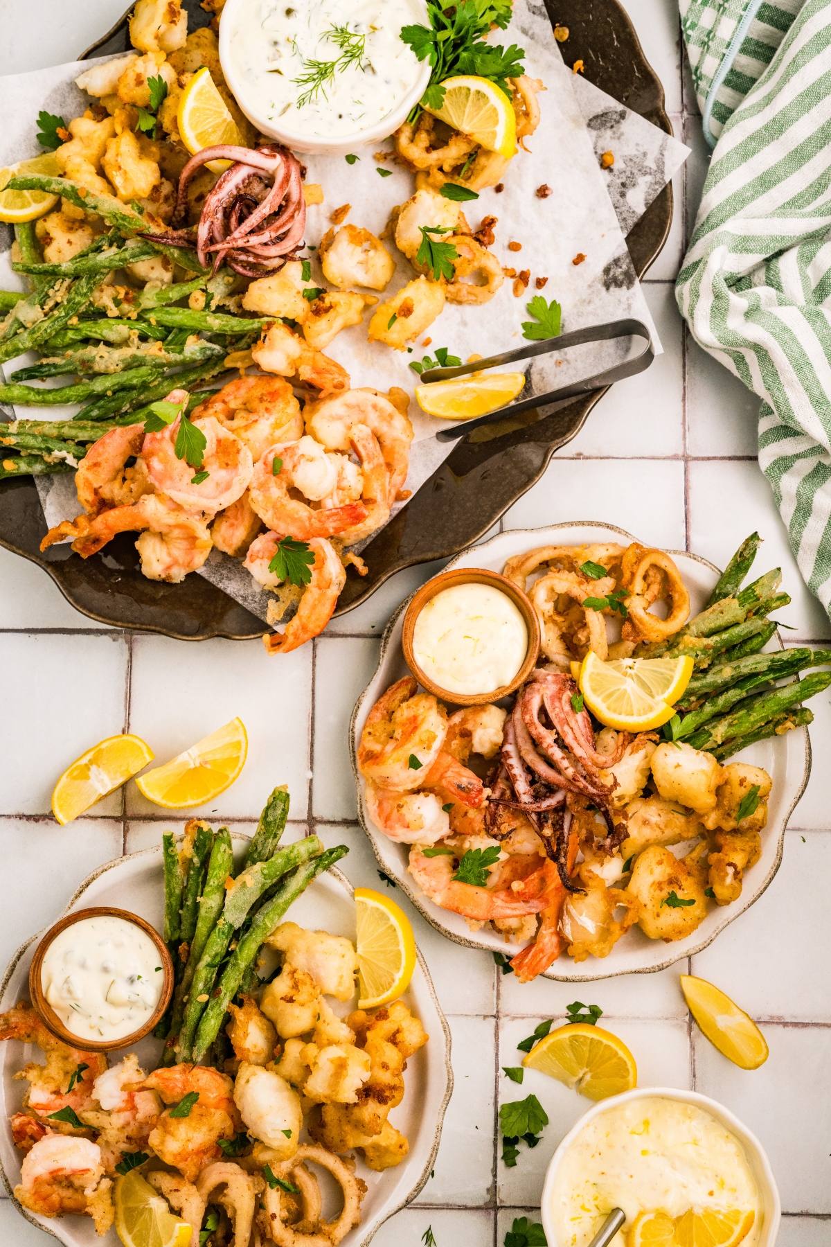 Plates of fried seafood, shrimp, green beans, lemon wedges, and dipping sauces on a white tiled surface.