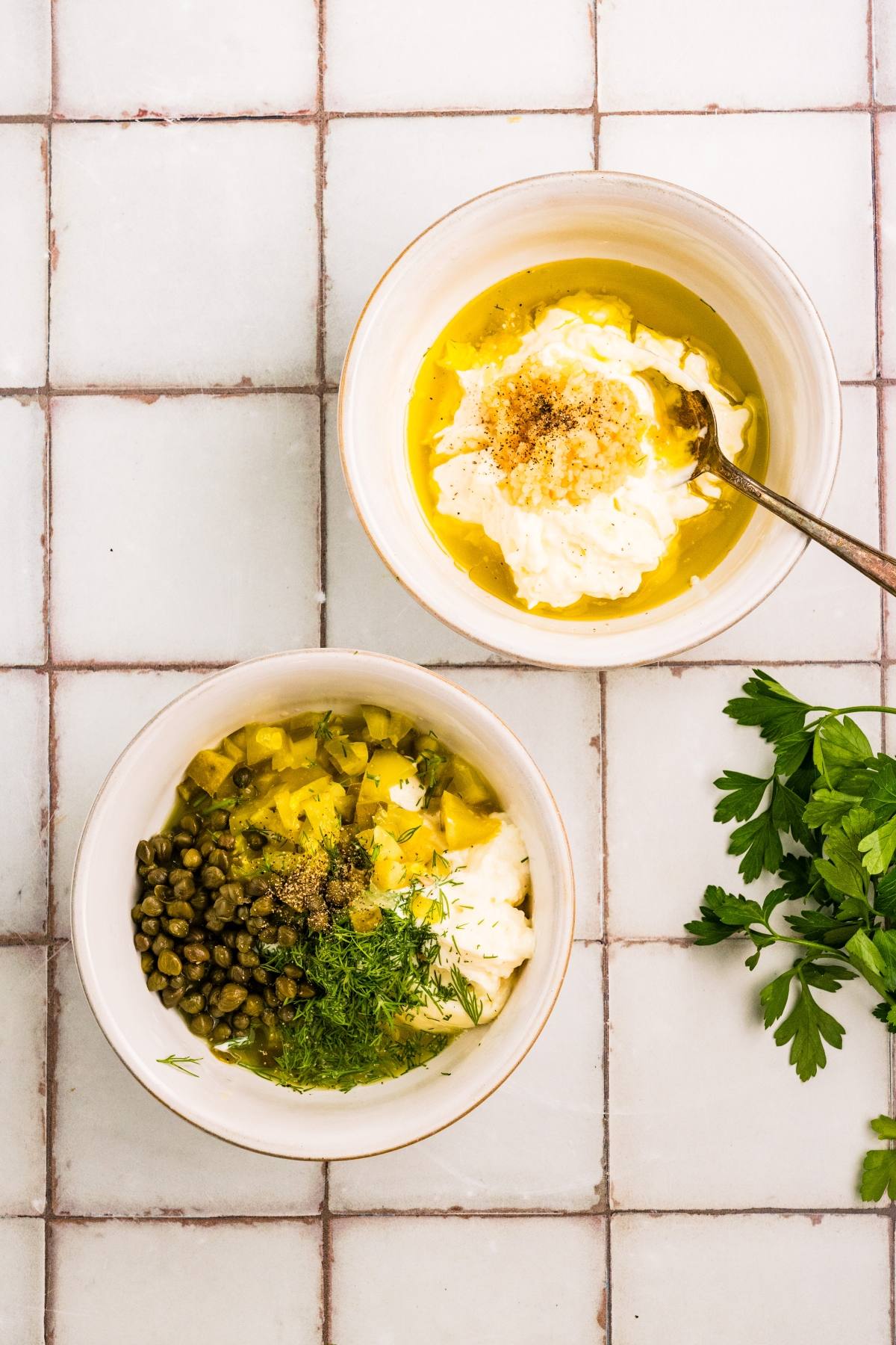 Two bowls on a tiled surface, one with creamy dip and oil, the other with capers, pickles, herbs, and sauce.