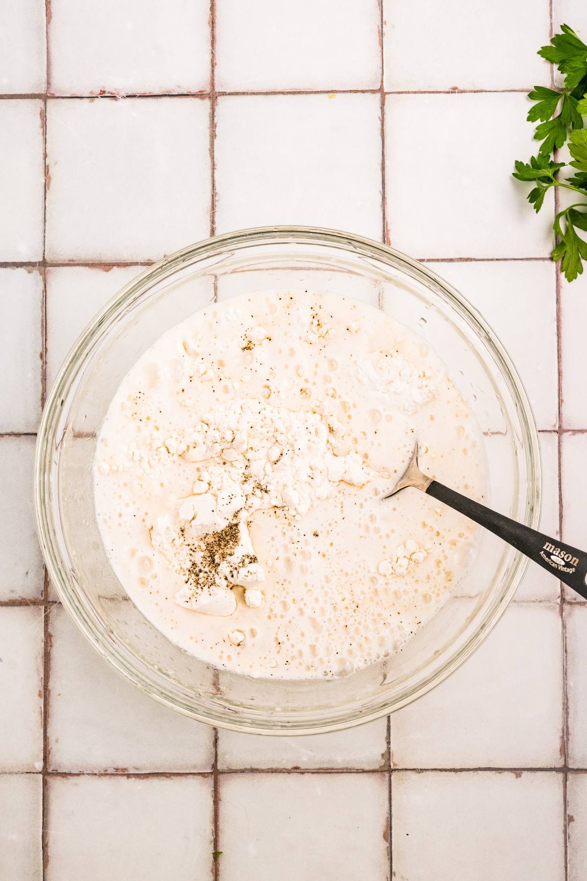 A glass bowl with flour, liquid, and pepper being mixed with a fork on a tiled surface.