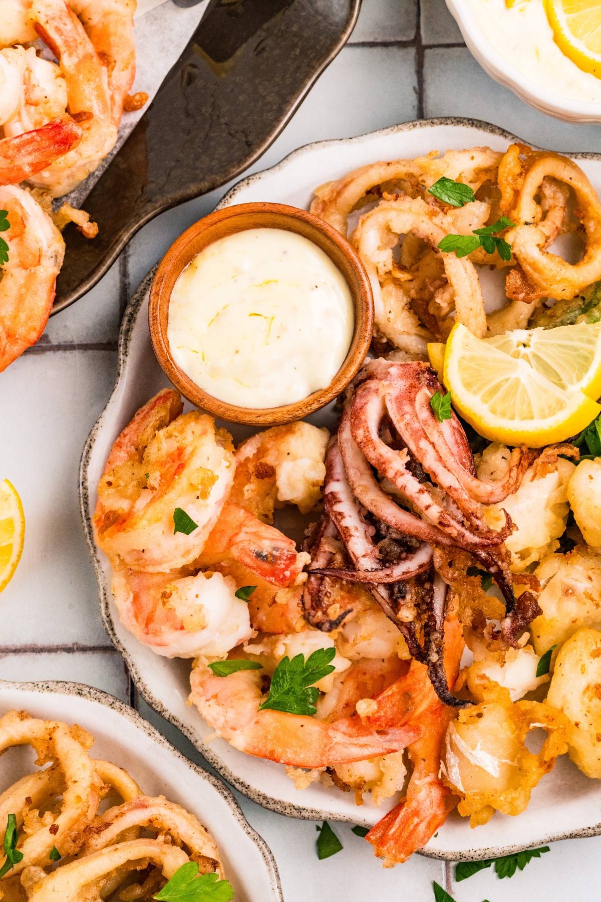 A plate of fried seafood including shrimp and calamari, garnished with parsley and lemon, with a sauce on the side.