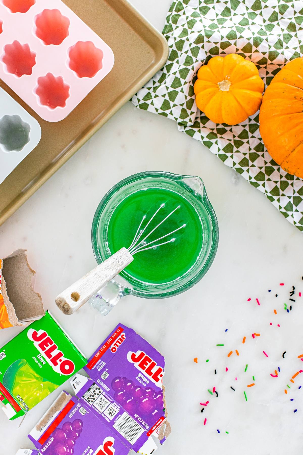 A bowl of green Jello mix with a whisk, Jello packets, pumpkins, and Halloween sprinkles on a table.