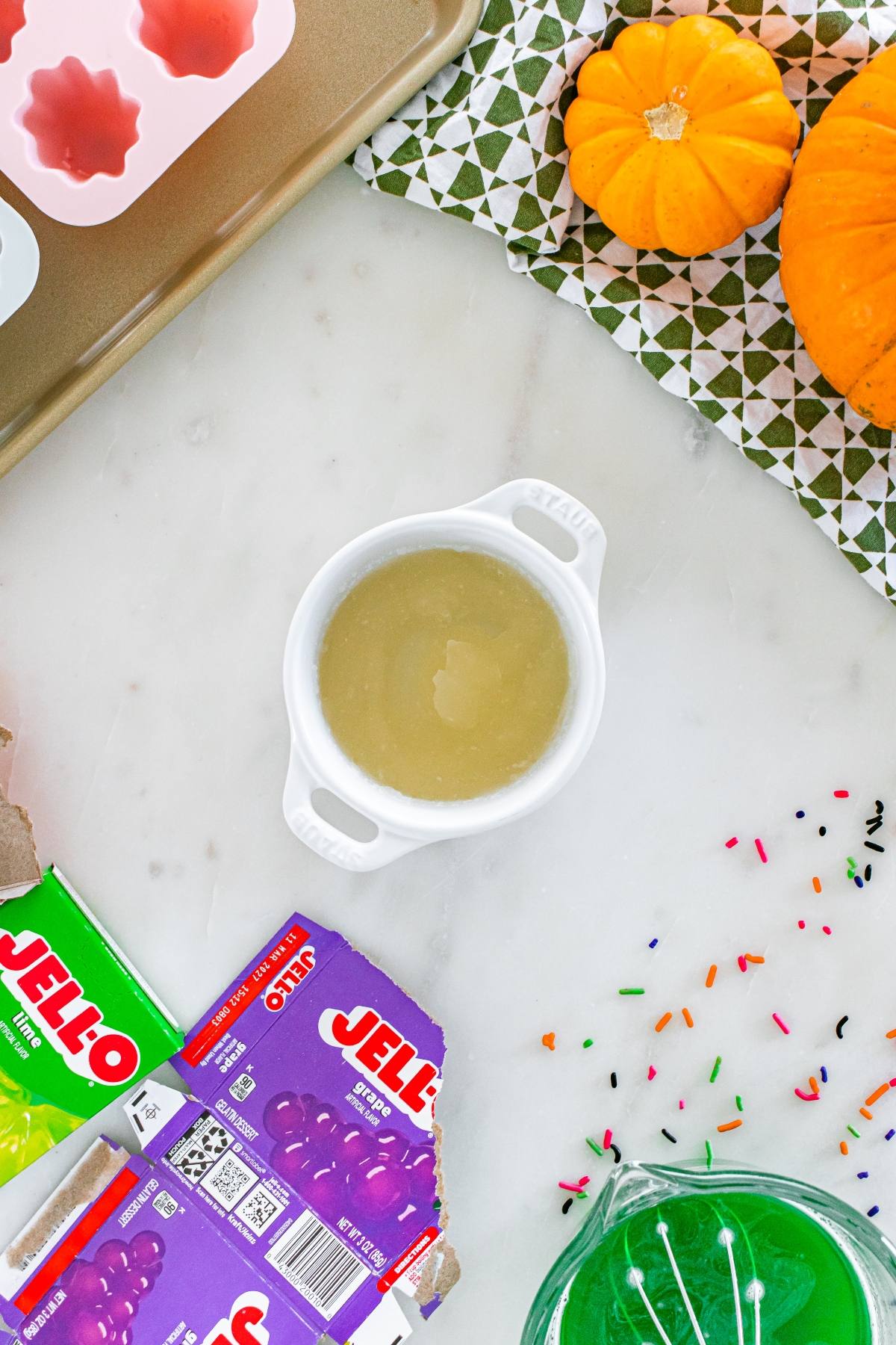 White bowl with liquid beside Jello boxes, sprinkles, pumpkins, and a green patterned cloth on a marble surface.