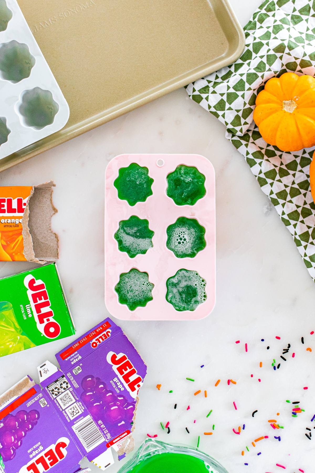 Green Jello in pumpkin-shaped silicone molds, surrounded by Jello boxes, a small pumpkin, and sprinkles.