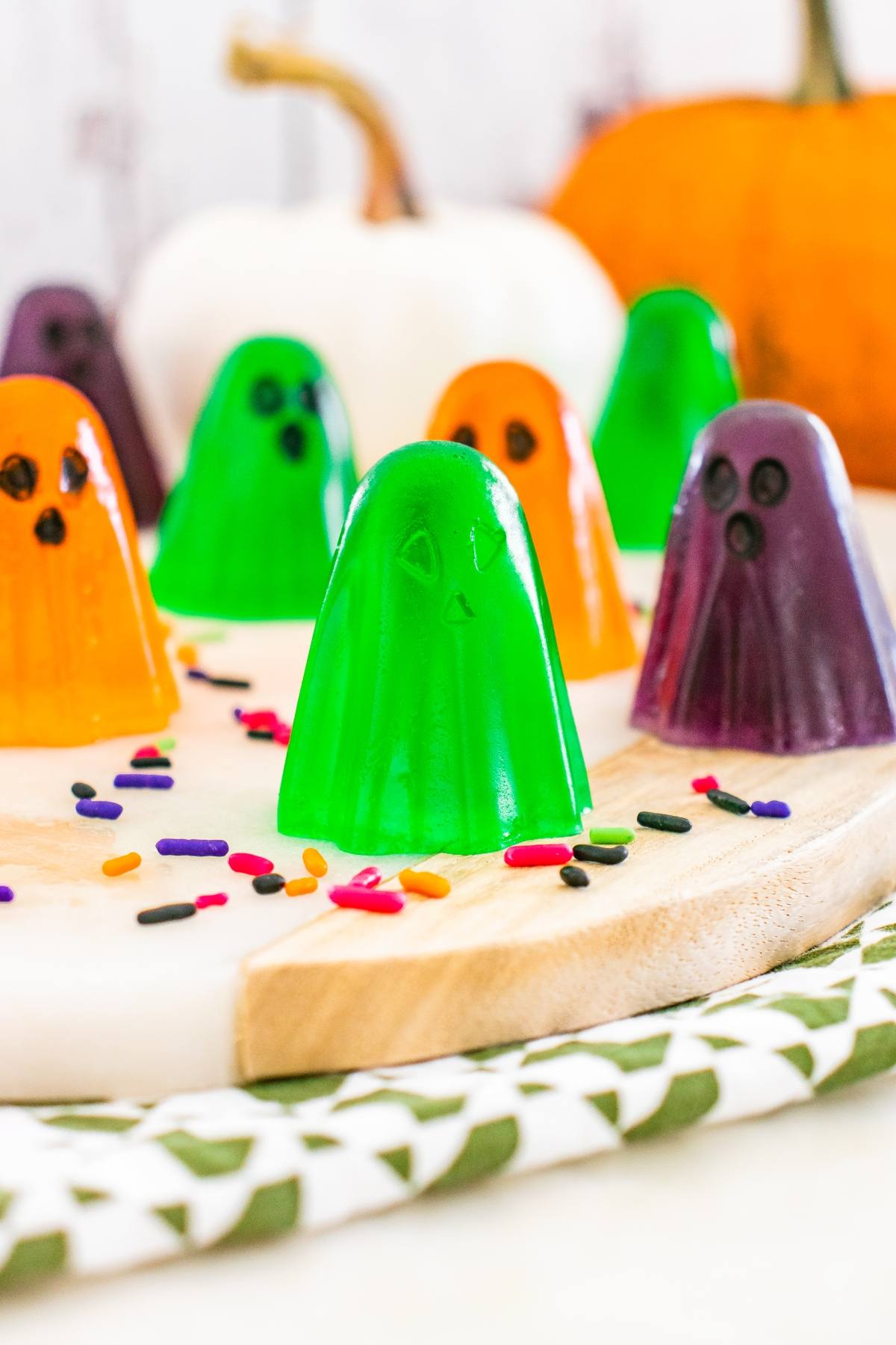 Colorful ghost-shaped gelatin candies with faces, surrounded by sprinkles and pumpkins in the background.