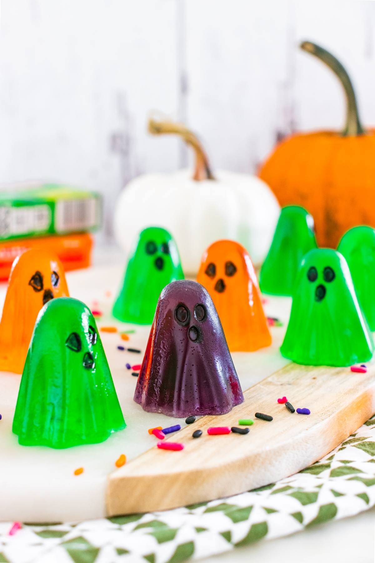 Colorful ghost-shaped gelatin snacks with candy faces on a cutting board, surrounded by sprinkles and pumpkins.