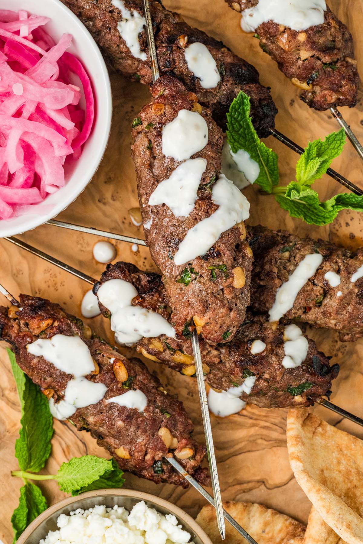 Beef kofta grilled kebabs with white sauce, fresh mint, pickled onions, pita bread, and feta cheese on a wooden board.