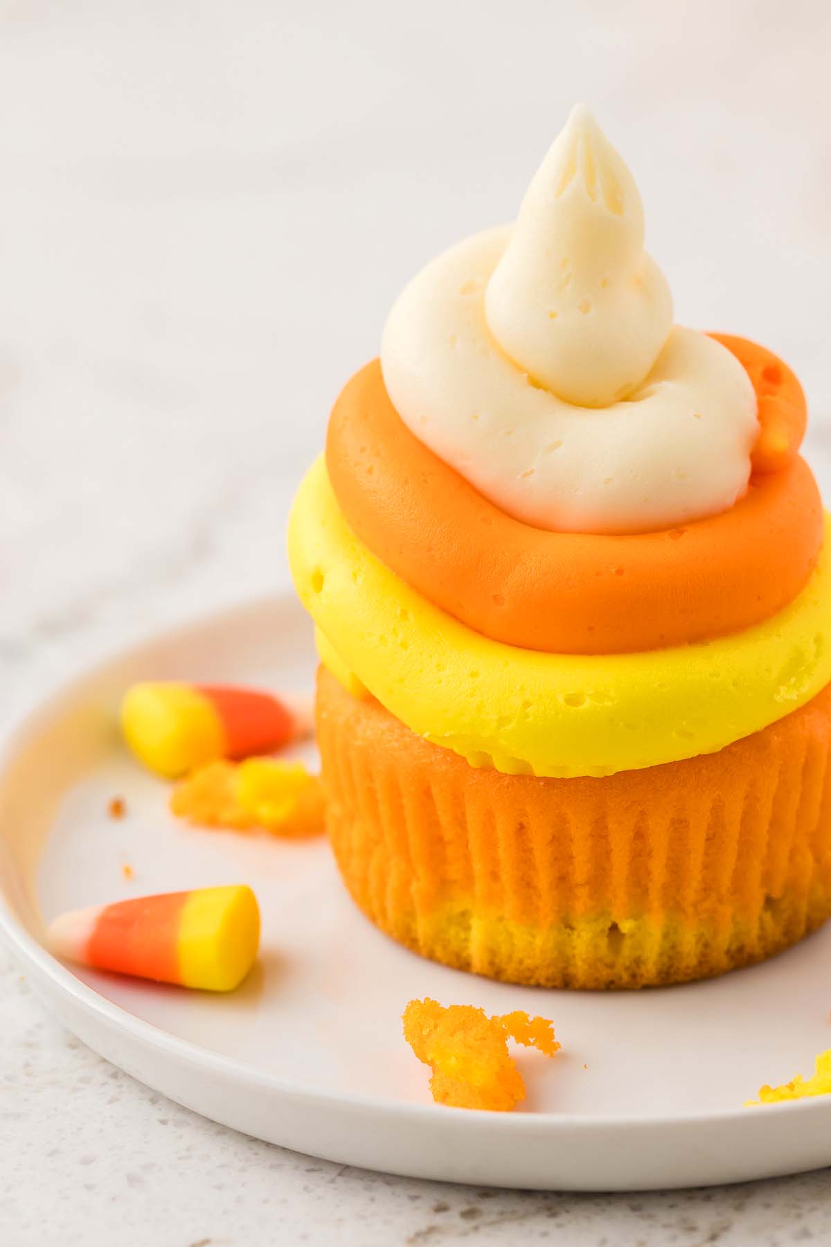 Candy corn cupcakes with yellow, orange, and white frosting, styled like classic candy corn, sit on a plate surrounded by festive candy corn pieces.