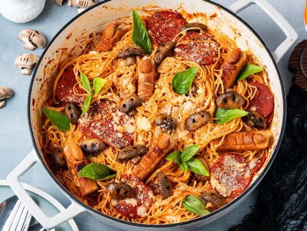 Halloween Pasta featuring spaghetti with tomato sauce, mushrooms, basil, and finger-shaped sausages in a white pot on the table.