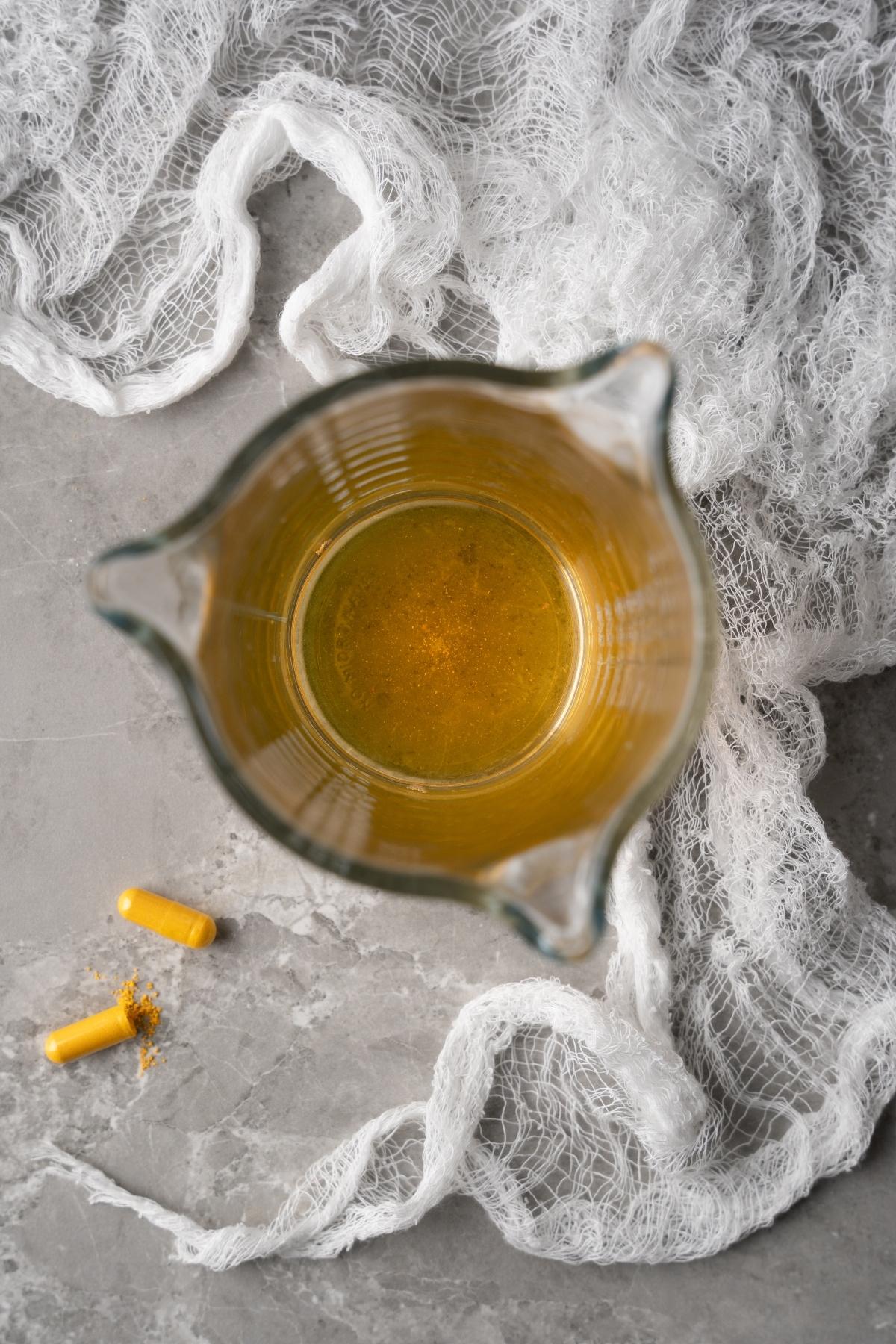 A glass pitcher with yellow liquid, resembling a fluorescent neon cocktail, sits beside two yellow capsules on a gray surface with white fabric.