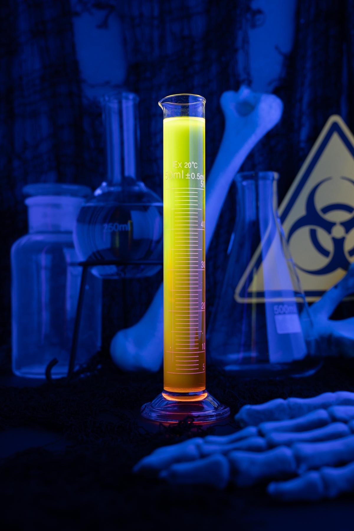 A glowing yellow-orange liquid, resembling a glow in the dark drink, sits in a graduated cylinder surrounded by lab glassware and bones.