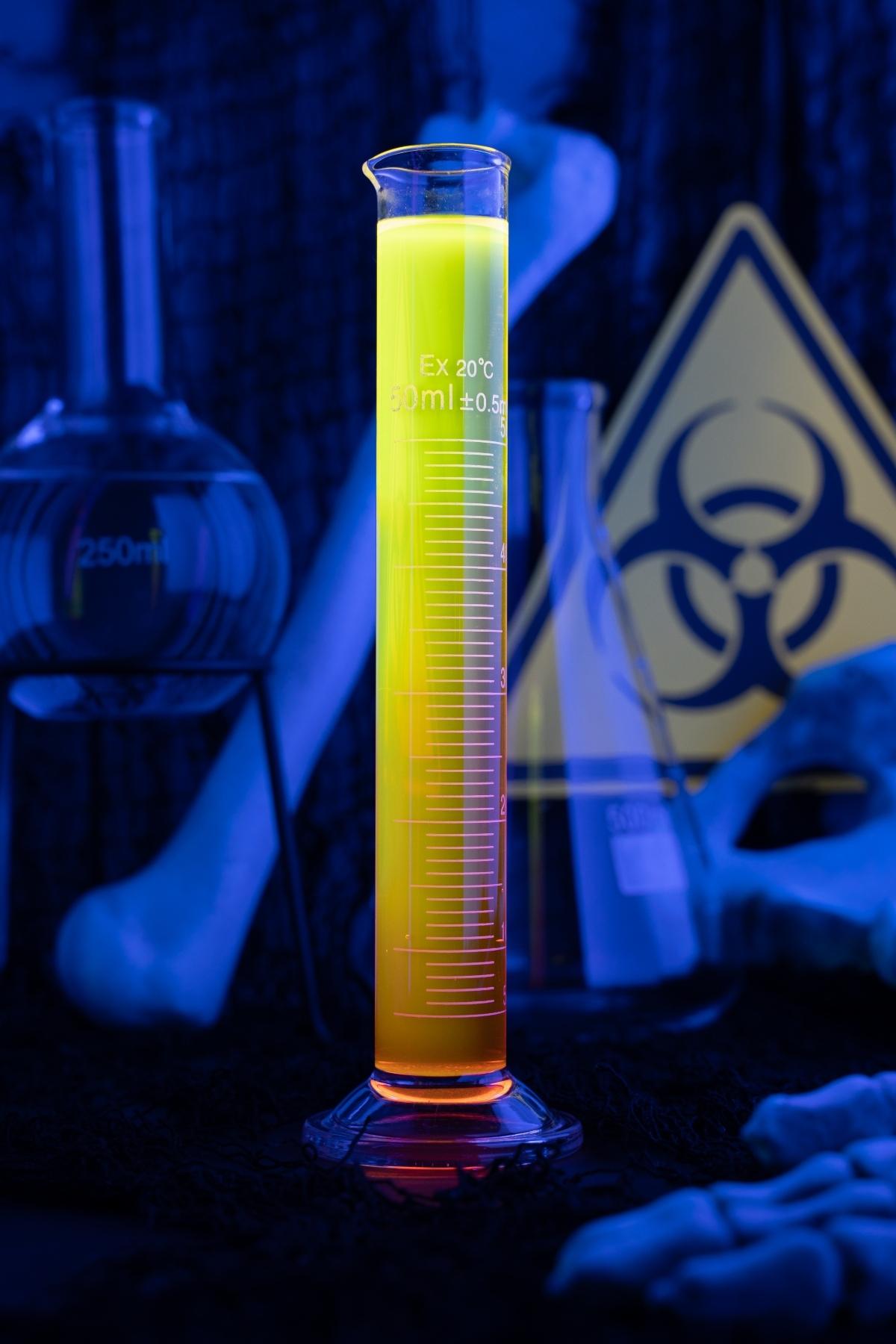 A glowing yellow liquid in a graduated cylinder, with lab glassware and a biohazard sign in the background—perfect for Halloween Glow in the Dark Shots or any fluorescent neon cocktail experiment.