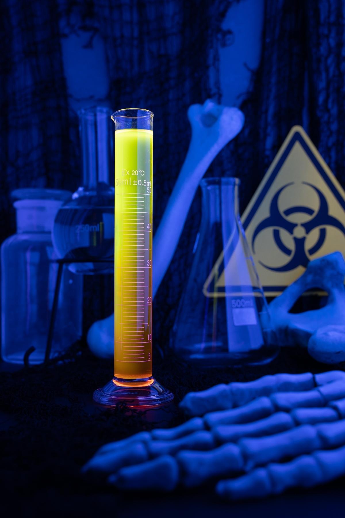 A glowing yellow liquid in a graduated cylinder with lab glassware, bones, and a biohazard sign in the background—perfect for Halloween Glow in the Dark Shots or showcasing vitamins that glow under black light.