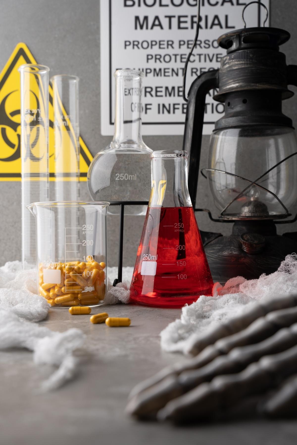 Laboratory glassware, yellow capsules resembling vitamins that glow under black light, and a lantern sit on a table with biohazard and warning signs in the background.