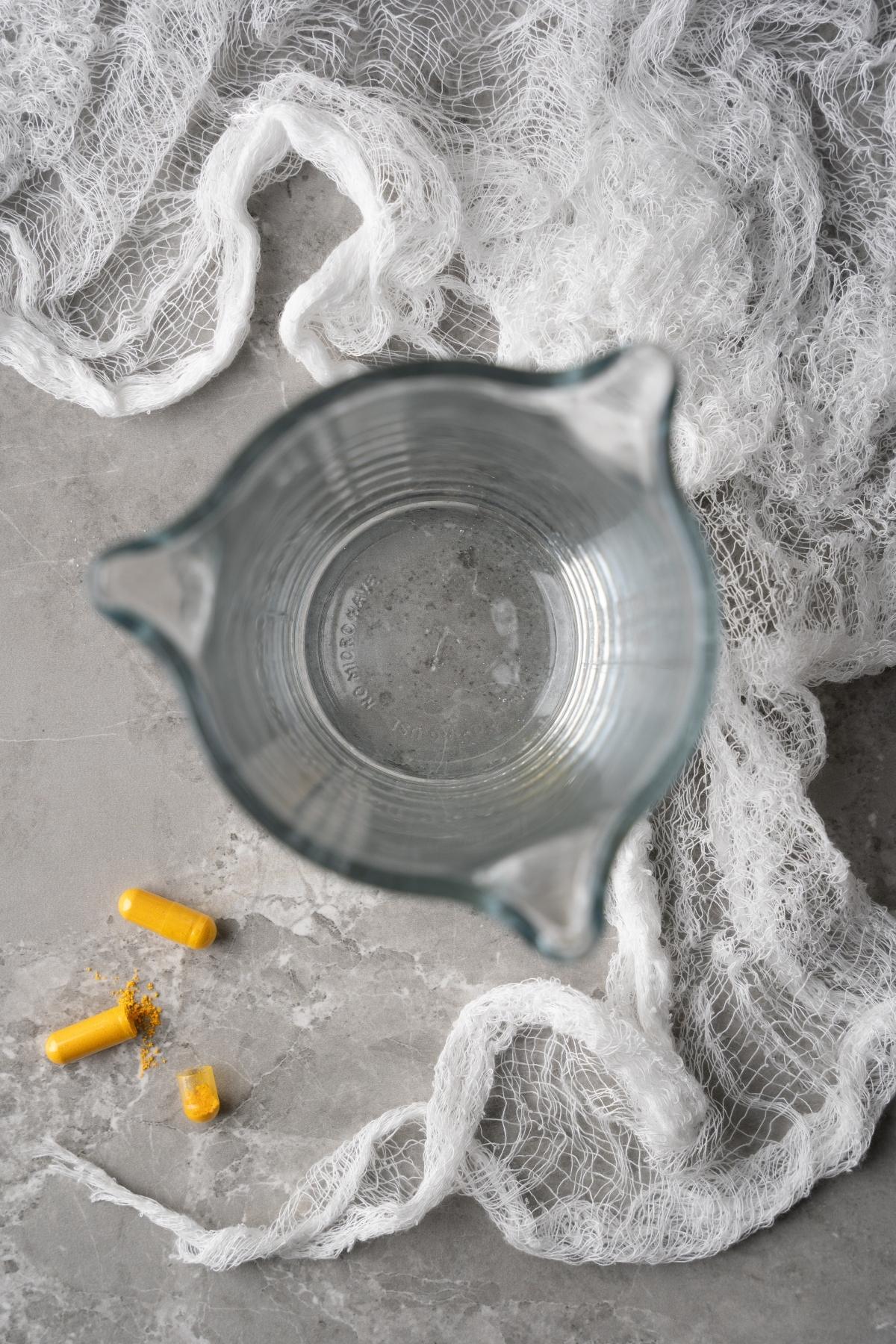 Glass of water seen from above, with two yellow capsules—resembling a glow in the dark drink—and white gauze on a gray surface.