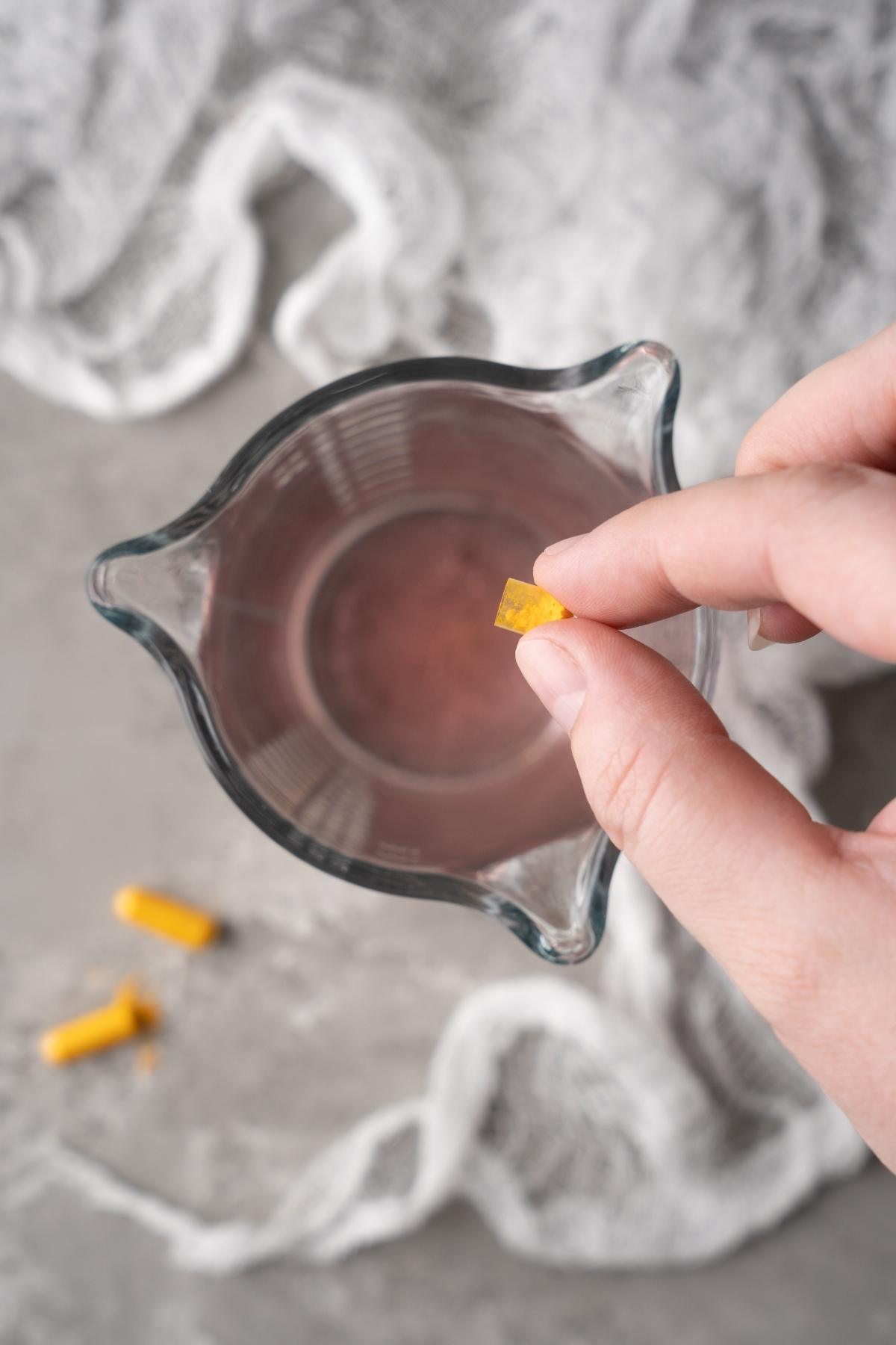 A hand drops a yellow tablet—like vitamins that glow under black light—into a glass of water on a gray surface.